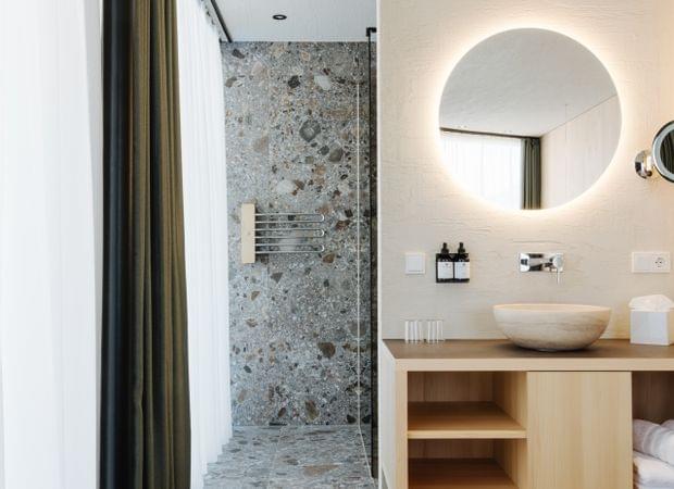 Moderner Badbereich mit rundem beleuchtetem Spiegel, Holzwaschtisch und offener Dusche mit steinverkleideter Wand.