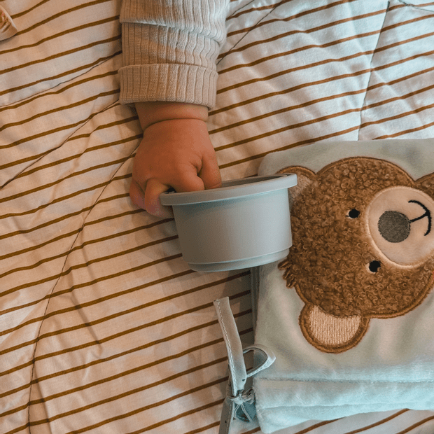 Babyhand hält grauen Becher auf gestreifter Decke neben Kuscheltuch mit Bärenmotiv.