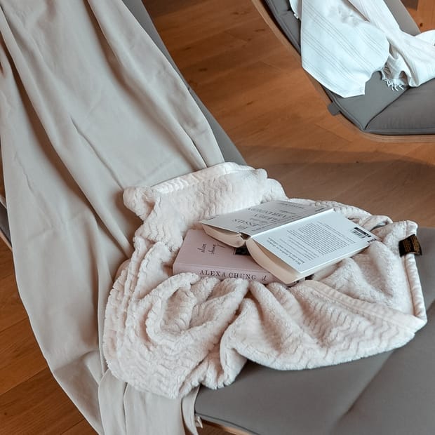 Liegestuhl mit beiger Auflage, darauf eine beige Decke, zwei Bücher, auf einem Holzboden.