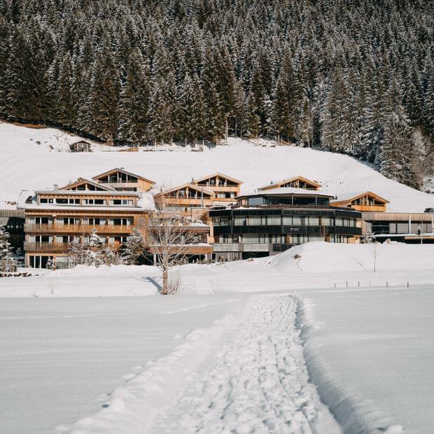 Verschneites alpines Hotelensemble, erreichbar über einen schmalen Pfad durch den Schnee, vor einem dichten Winterwald