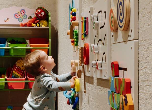 Kleines Kind spielt an einer bunten Aktivwand mit drehbaren und verschiebbaren Elementen neben einem Regal voller Spielzeug.