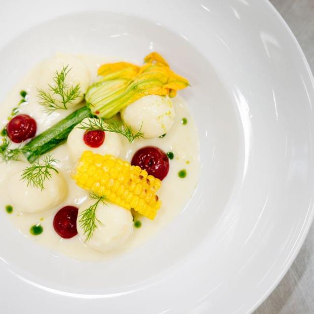Fein angerichtetes Gourmetgericht auf weißem Teller mit cremigen Elementen, Früchten, Kräutern und essbaren Blüten