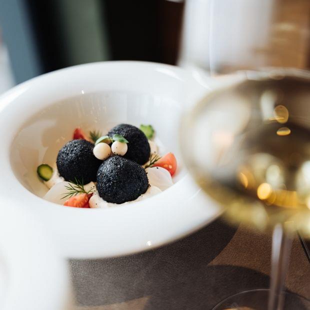 Dessert mit Beeren und cremigen Elementen in einer weißen Schale, daneben ein Glas Weißwein auf gedecktem Tisch