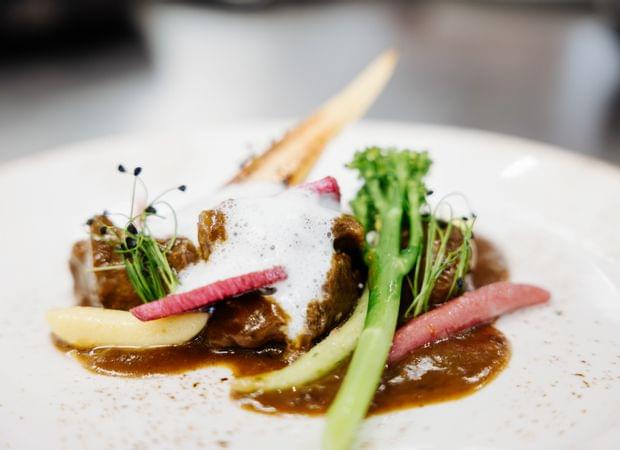 Detailaufnahme eines kunstvoll angerichteten Gourmetgerichts mit Fleisch, Sauce, grünem Gemüse und Kräutern auf weißem Teller.