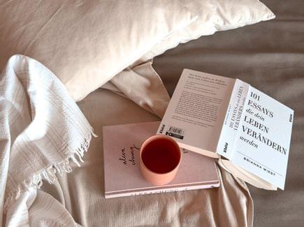 Gemütliche Liege mit beigen Kissen und Decke, darauf ein geöffnetes Buch, ein rosa Buch und eine Tasse.