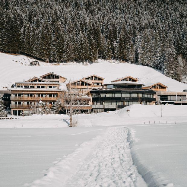 Verschneites alpines Hotelensemble, erreichbar über einen schmalen Pfad durch den Schnee, vor einem dichten Winterwald