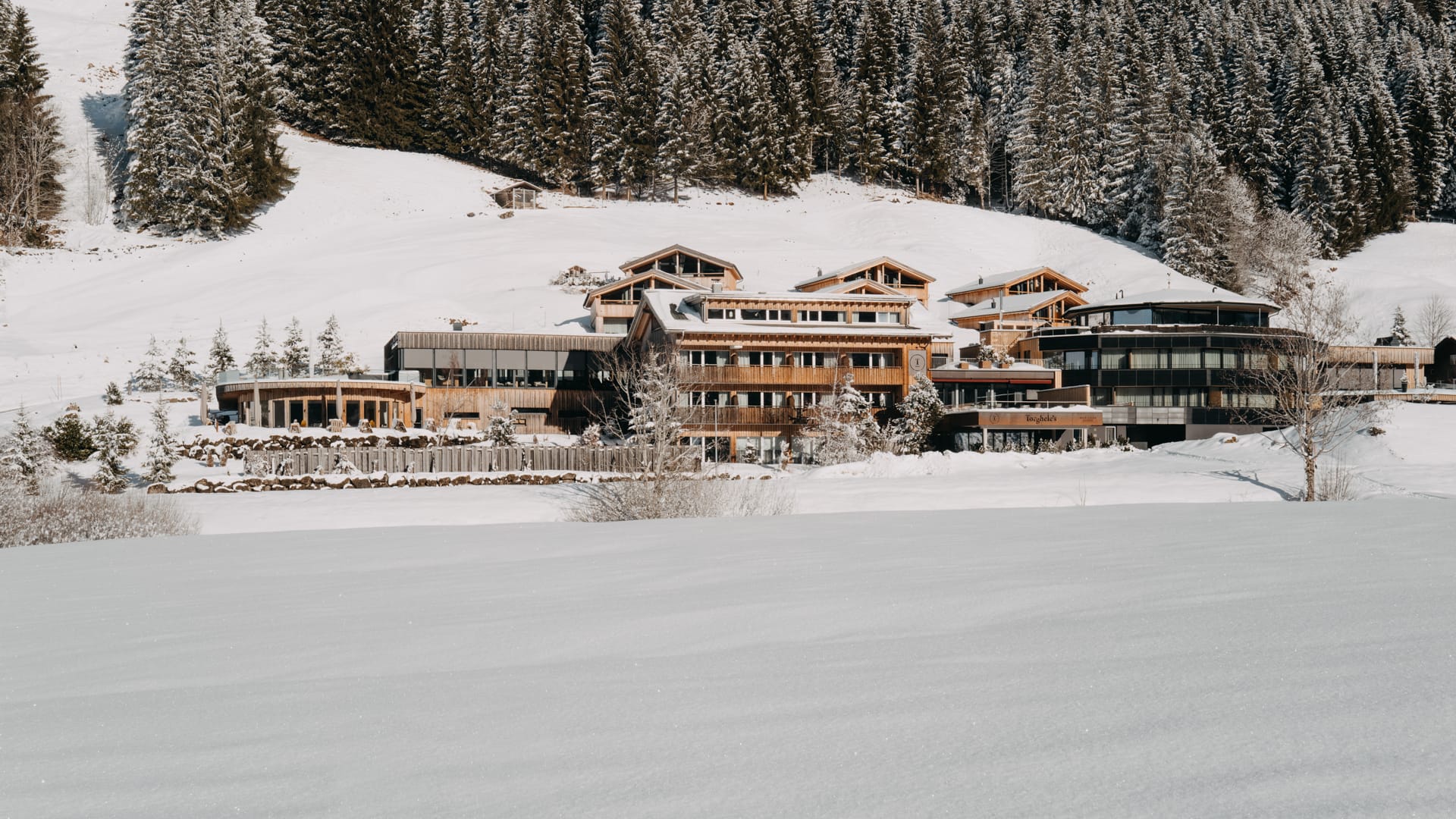Alpines Hotelensemble auf einer offenen, schneebedeckten Fläche, im Hintergrund ein dichter Winterwald.