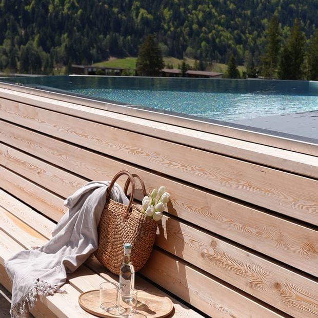 Holzterrasse mit geflochtenem Korb, Tulpen und Getränken neben einem Infinity-Pool vor bewaldeter Bergkulisse.