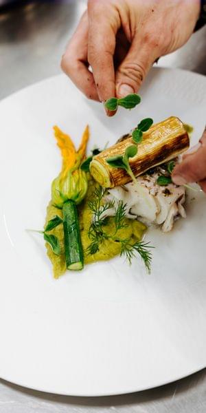 Hände richten ein kunstvoll angerichtetes Gourmetgericht auf einem weißen Teller mit Kräutern und Gemüse an