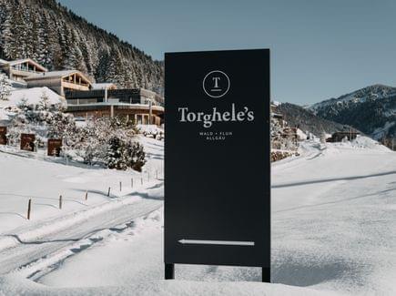 Schwarzes Hotelschild im Schnee vor einem verschneiten alpinen Hotelensemble und winterlicher Berglandschaft