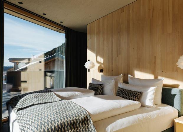 Modernes Doppelzimmer mit Holzverkleidung, großen Fenstern, weißer Bettwäsche und gemusterten Decken und Kissen.