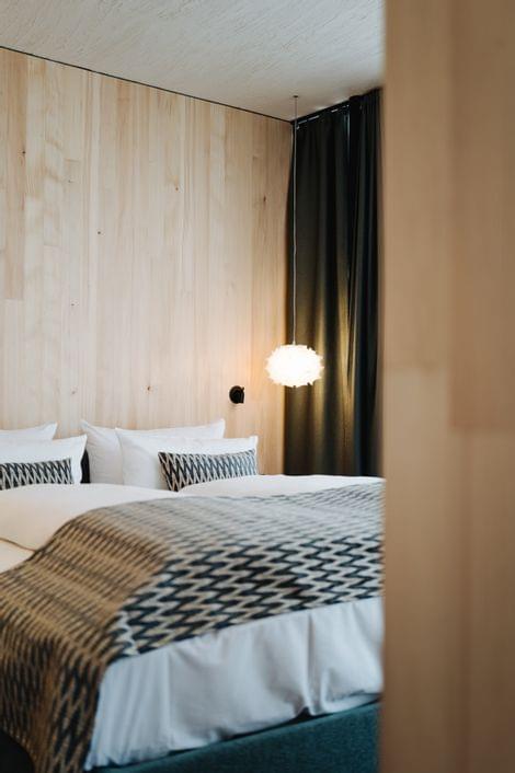 Modernes Schlafzimmer mit Holzverkleidung, großem Bett mit gemustertem Überwurf und Kissen sowie einer weißen Hängelampe neben dunklen Vorhängen.