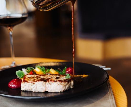 Hände gießen Sauce aus einer Kanne über ein kunstvoll angerichtetes Gourmetgericht auf schwarzem Teller