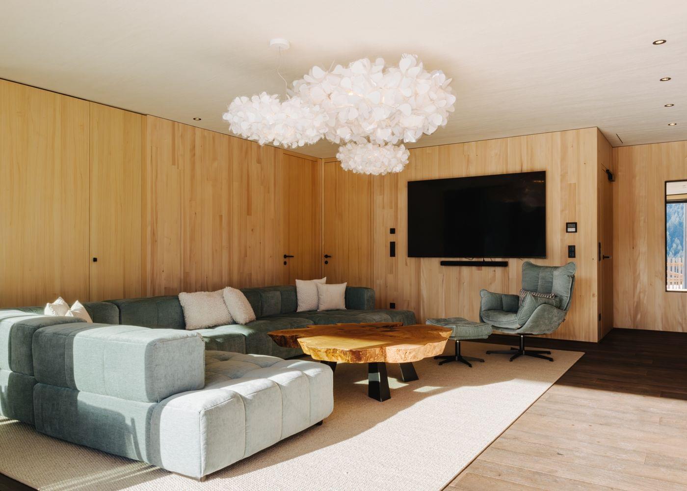 Geräumiger Wohnbereich mit heller Holzverkleidung, großer Ecksofa-Landschaft, rundem Holztisch, gemütlichem Sessel und dekorativer Deckenleuchte.