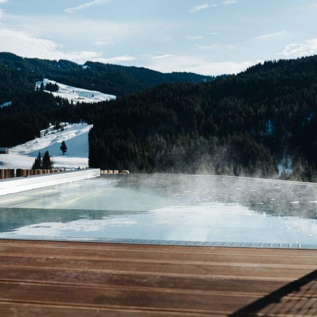 Beheizter Außenpool mit aufsteigendem Dampf und Blick auf verschneite Berghänge und Wälder unter blauem Himmel.