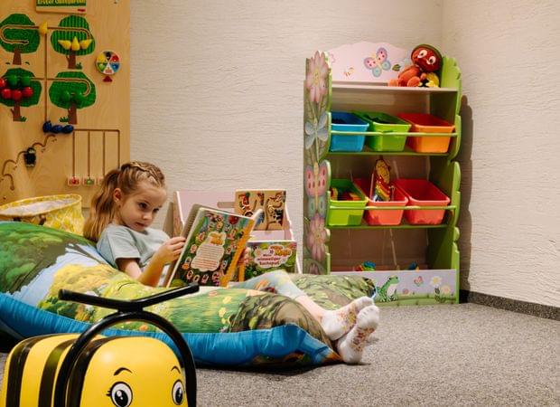 Kind sitzt entspannt auf großen bunten Kissen und schaut ein Bilderbuch, daneben ein farbenfrohes Regal mit Spielzeug.