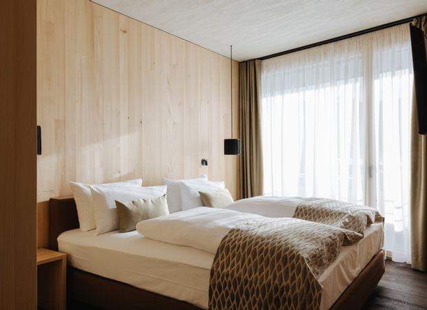 Gemütliches Schlafzimmer mit großem Doppelbett, hellen Holzwänden, weißen Kissen und gemustertem Überwurf vor einer bodentiefen Fensterfront.