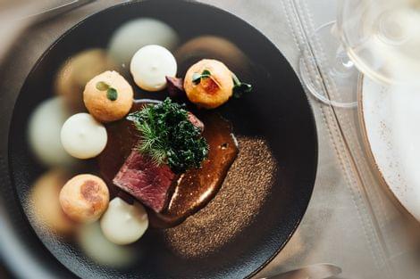 Fein angerichtetes Gourmetgericht auf schwarzem Teller mit Fleisch, kleinen Beilagen und Kräutern, von oben fotografiert