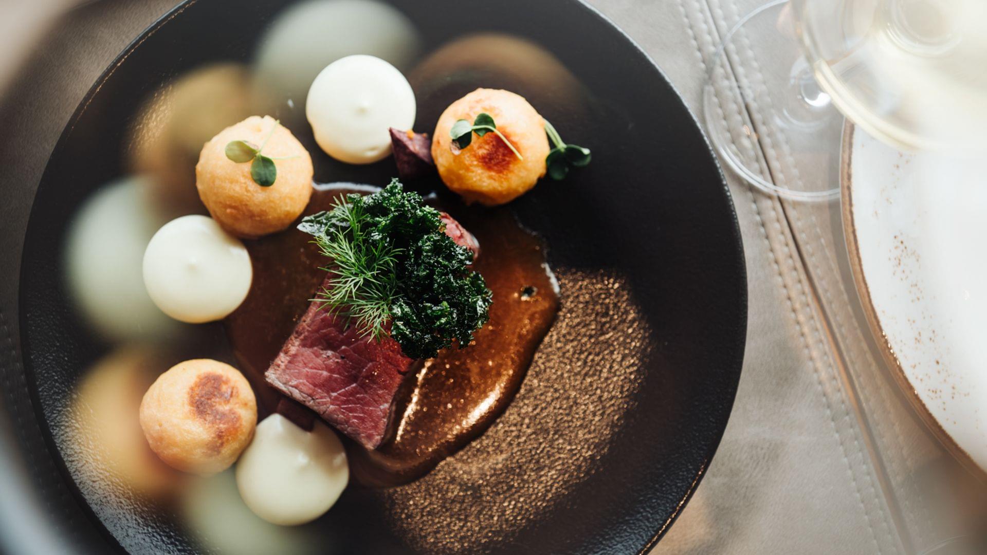 Fein angerichtetes Gourmetgericht auf schwarzem Teller mit Fleisch, kleinen Beilagen und Kräutern, von oben fotografiert