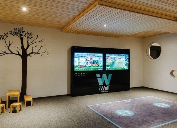 Kinderbereich mit digitaler Spielwand, kleinem Holztisch mit Hockern, Wanddekor in Baumform und beleuchtetem Spielteppich am Boden.