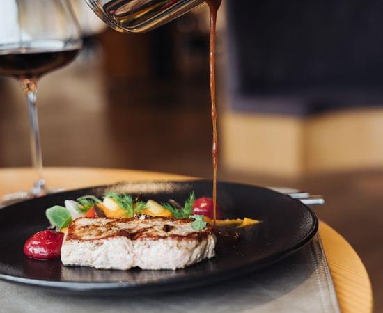 Hände gießen Sauce aus einer Kanne über ein kunstvoll angerichtetes Gourmetgericht auf schwarzem Teller