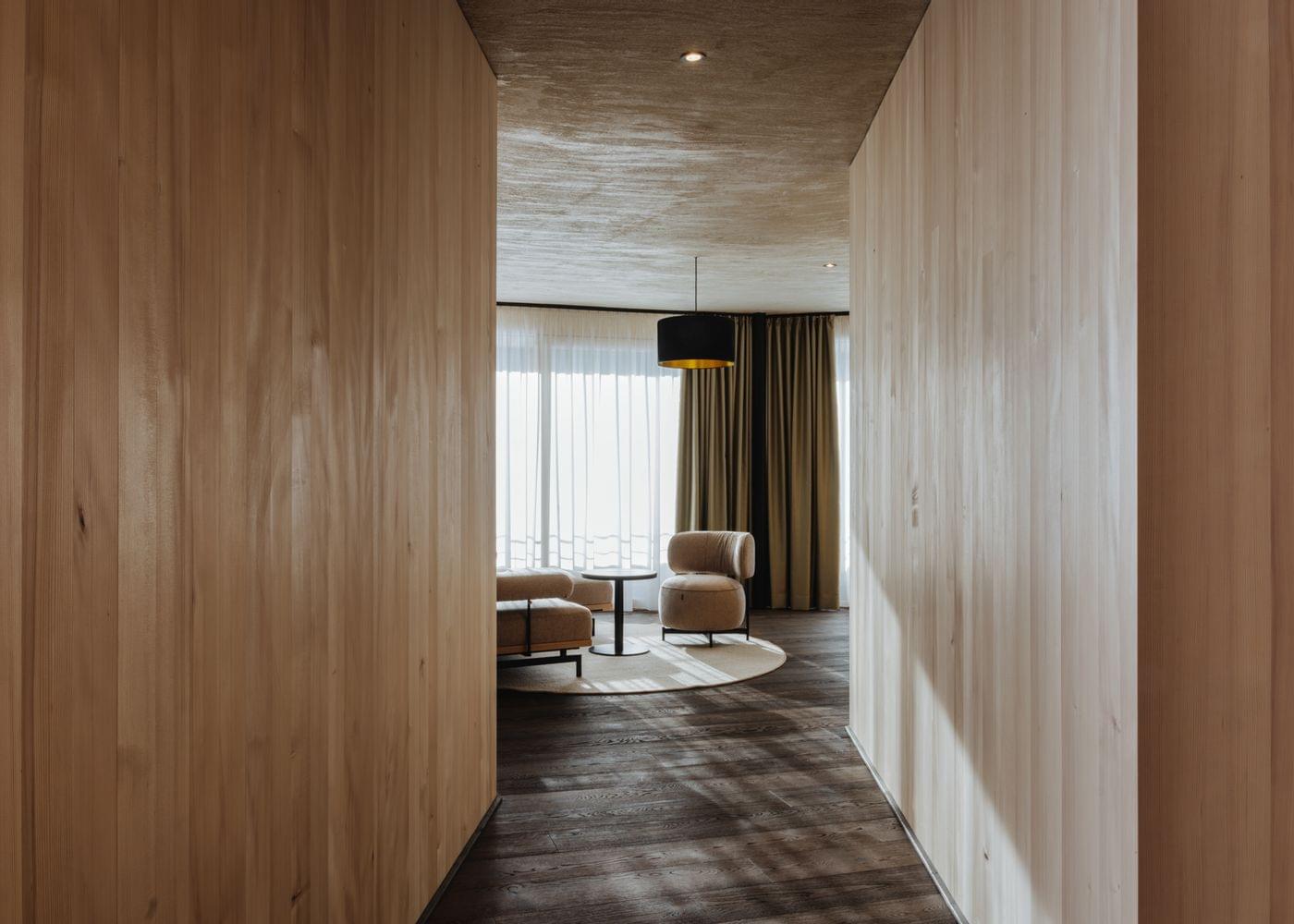 Holzverkleideter Flur mit Blick in einen hellen Wohnbereich mit Sesseln und großen Fenstern.